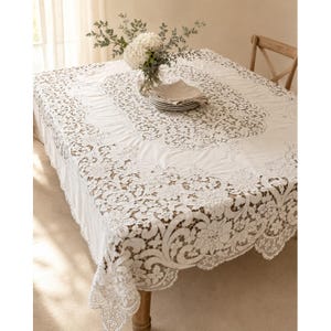May include: A white rectangular tablecloth featuring an intricate lace border with floral and scroll patterns. The center of the fabric is solid white, and the lace detailing extends along the edges, draping over the sides of the table.