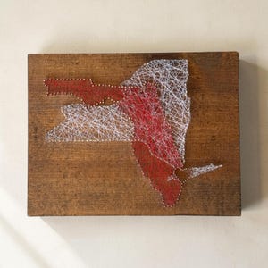 May include: A rectangular wooden plaque featuring a string art design of the state of New York. The map outline is created with small metal nails and interwoven white and red thread, creating a dense, layered geometric pattern on the dark wood.