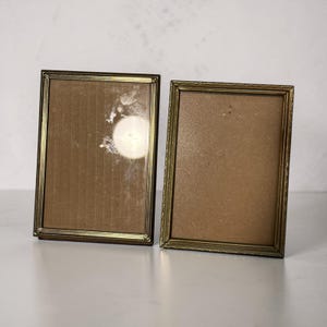 May include: Two rectangular picture frames with metallic gold-toned borders featuring a fine beaded texture along the edges. Each frame contains a plain brown backing board, and the left frame has a clear glass front with visible light reflections.