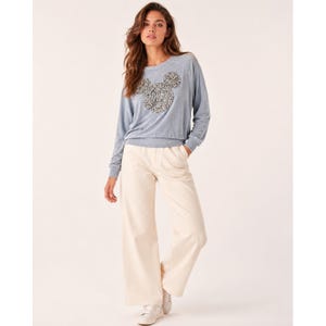 Puede incluir: Una sudadera de manga larga en color azul claro con un gráfico central de una silueta de cabeza de ratón rellena de lentejuelas plateadas, combinada con pantalones anchos de color crema y zapatillas blancas. El conjunto es casual y tiene cuello redondo.