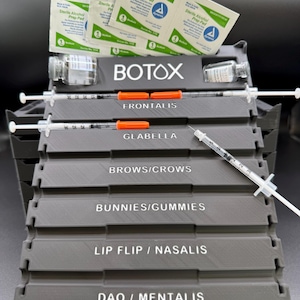 May include: A gray tiered organizer with the word "BOTOX" in white. It holds syringes, vials, and alcohol prep pads. The organizer is labeled with treatment areas: Frontalis, Glabella, Brows/Crows, Bunnies/Gummies, Lip Flip/Nasalis, and DAO/Mentalis.