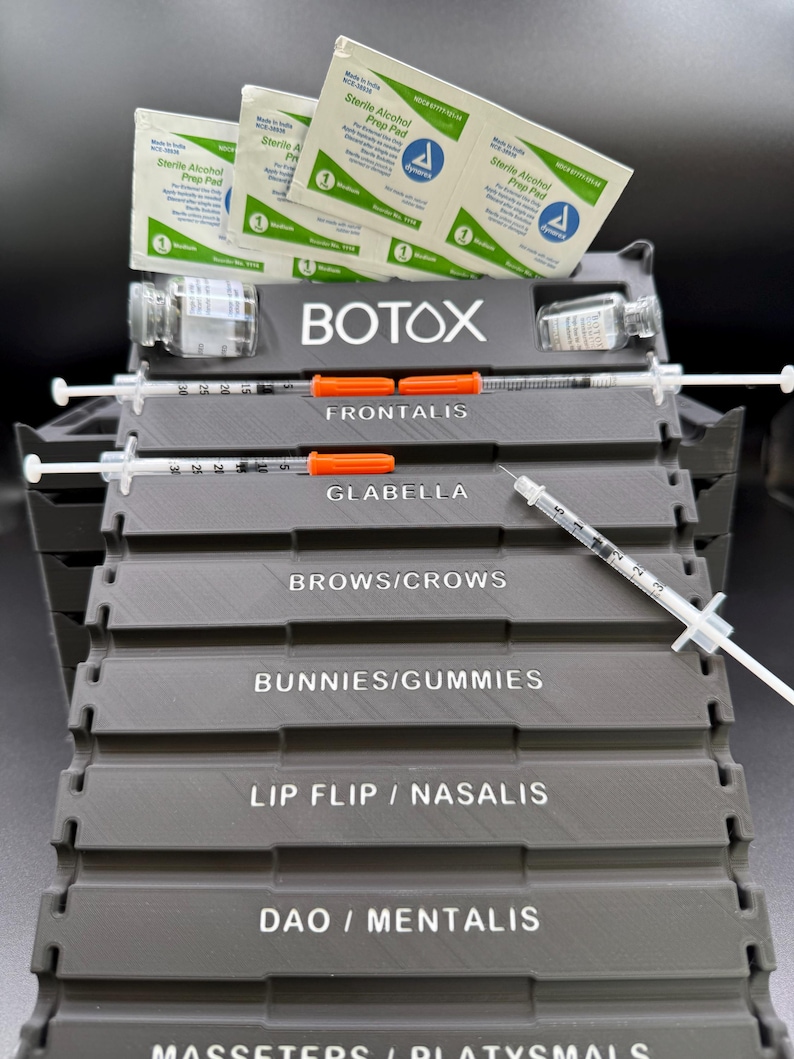 May include: A gray tiered organizer with the word "BOTOX" in white. It holds syringes, vials, and alcohol prep pads. The organizer is labeled with treatment areas: Frontalis, Glabella, Brows/Crows, Bunnies/Gummies, Lip Flip/Nasalis, and DAO/Mentalis.