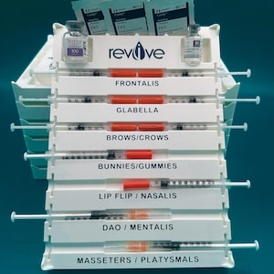May include: A white plastic tray with compartments for facial treatments, containing syringes, vials of Botox, and small packets. The tray is labelled with the word "revive". Compartments are labelled: Frontalis, Glabella, Brows/Crows, Bunnies/Gummies, Lip Flip/Nasalis, Dao/Mentalis, and Masseters/Platysmals.