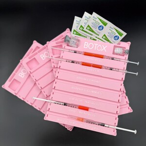 May include: Pink plastic trays with the word "BOTOX" printed on them. Each tray has slots for syringes and vials. Syringes are filled with an orange liquid. Alcohol prep pads and vials are also visible.