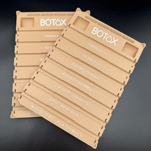 May include: Two beige plastic organizers with the word "BOTOX" in white. Each organizer has labeled sections for facial areas like "Frontalis" and "Glabella". The organizers are designed to hold items related to cosmetic procedures.