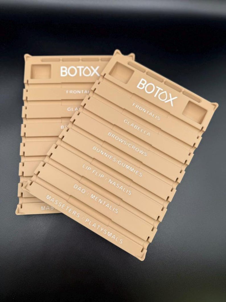 May include: Two beige plastic organizers with the word "BOTOX" in white. Each organizer has labeled sections for facial areas like "Frontalis" and "Glabella". The organizers are designed to hold items related to cosmetic procedures.
