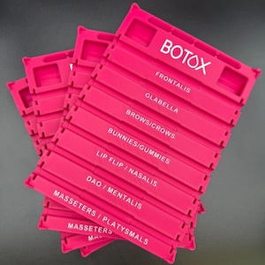 May include: Several bright pink plastic organizers are stacked on a dark surface. Each organizer has multiple labeled sections with white text, including "BOTOX," "FRONTALIS," and "GLABELLA."