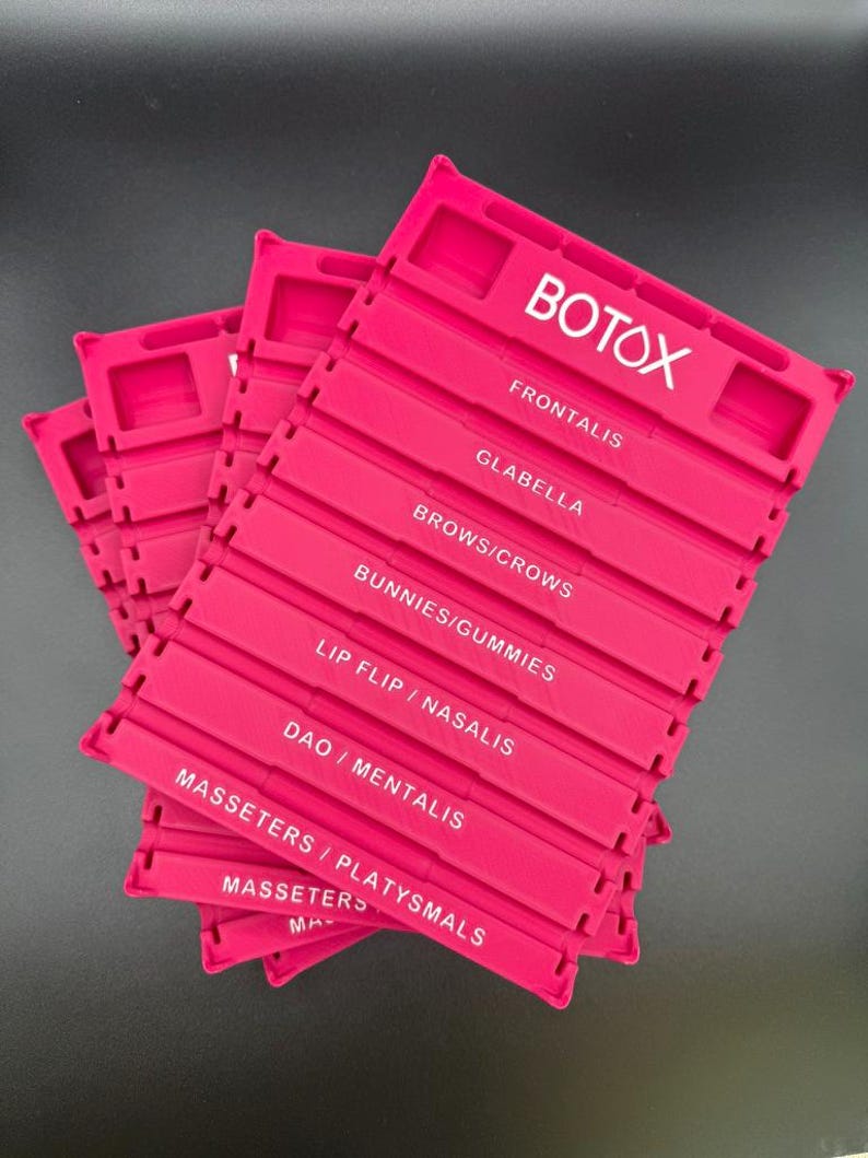 May include: Several bright pink plastic organizers are stacked on a dark surface. Each organizer has multiple labeled sections with white text, including "BOTOX," "FRONTALIS," and "GLABELLA."