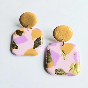 Puede incluir: Un par de pendientes hechos a mano. Cada pendiente presenta una base cuadrada rosa claro con detalles de pan de oro y formas de color lavanda y mostaza. Un perno circular de color mostaza remata cada pendiente.