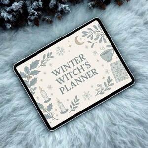 May include: A digital tablet displaying a cream-colored planner design with the text "Winter Witch's Planner." The design includes illustrations of holly, snowflakes, candles, a teacup, a crescent moon, and a tarot card. The tablet is set on a fluffy, light blue surface.