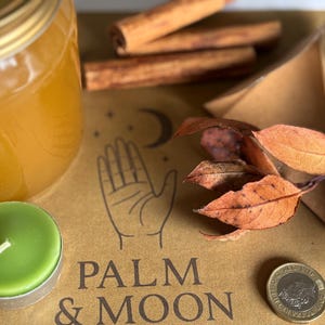 May include: An arrangement featuring a jar of honey, a green tea light candle, cinnamon sticks, dried leaves, a pen, and a coin. The background includes the text "PALM & MOON" and a hand illustration.