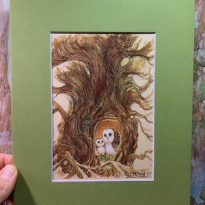 May include: Watercolor painting of two owls in a tree hollow, framed with a green mat. The artwork features a detailed depiction of the tree's textured bark and the owls' soft feathers. Signed "J. Brown '23".