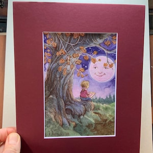 May include: Watercolor painting of a child reading under a tree at night, with a smiling moon in the background. The artwork is framed in a burgundy mat.