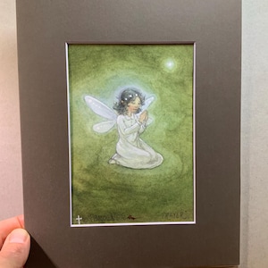 May include: A framed watercolor painting of a kneeling fairy with white wings and a halo. The fairy is wearing a white dress and has dark hair. The background is green with a bright light. The painting is titled "Prayer".