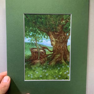 May include: A framed watercolor painting depicts a whimsical scene of a tree with a face, sheltering a smaller tree and a wooden bucket. The artwork is set in a green field with small white flowers, under a blue sky. The painting is matted in a green frame.