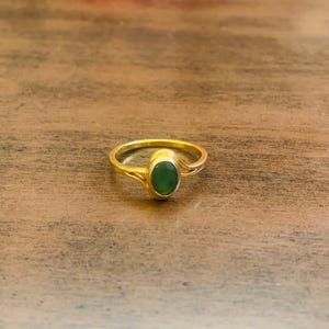May include: A gold ring featuring an oval, emerald-green gemstone. The ring is displayed on a wooden surface. The ring's band has a simple, elegant design. The gemstone is the central feature of the ring.
