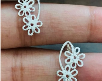 May include: A pair of silver-colored earrings featuring a floral design. Each earring has two openwork flowers connected by a curved stem. The earrings are shown against a blurred background.