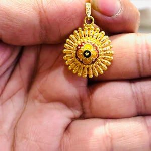 May include: A circular gold pendant featuring a sunburst design with radiating textured rays. The center of the pendant displays a floral motif with red, yellow, and black enamel detailing. A small gold loop is attached to the top for hanging.