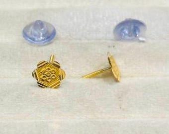 May include: Two gold-colored flower-shaped stud earrings with intricate detailing, alongside two clear, rubber earring backs. The earrings are displayed on a white, textured surface.