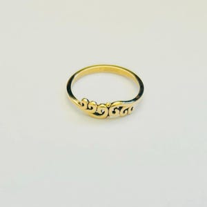 Op de afbeelding: Een goudkleurige ring met een sierlijk, wervelend ontwerp aan de voorkant. De ring staat tegen een witte achtergrond, wat de ingewikkelde details en de gepolijste afwerking benadrukt. De ring heeft een dunne en delicate band.