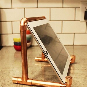 Pode incluir: Um suporte para tablet feito de tubos de cobre com um tablet branco. O suporte é feito de tubos e conexões de cobre, com uma base em forma de T e um suporte vertical. O tablet está inclinado para visualização.