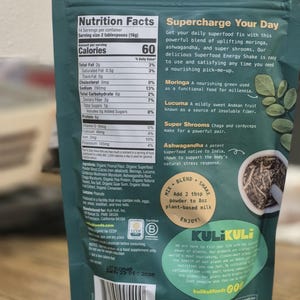 Op de afbeelding: Een groene en turquoise zak van Kuli Kuli Superfood Energy Shake. De verpakking toont voedingsinformatie, ingredi&euml;ntgegevens en de tekst "Supercharge Your Day". Het bevat ook een "Mix, Blend, Shake" instructie.