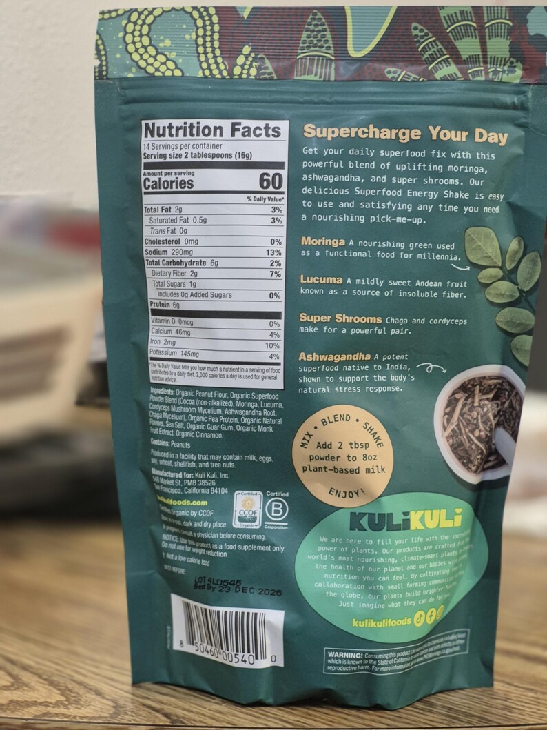 Op de afbeelding: Een groene en turquoise zak van Kuli Kuli Superfood Energy Shake. De verpakking toont voedingsinformatie, ingredi&euml;ntgegevens en de tekst "Supercharge Your Day". Het bevat ook een "Mix, Blend, Shake" instructie.