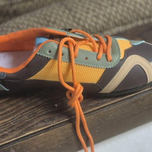 Könnte beinhalten: Ein farbenfroher Sportschuh mit orangefarbenen Schnürsenkeln, einem gelben Mesh-Einsatz und braunen und beigen Akzenten. Der Schuh hat ein gepolstertes orangefarbenes Innenfutter und eine hellblaue Zierleiste. Der Schuh ist auf Komfort und Stil ausgelegt.