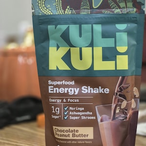 Op de afbeelding: Een verpakking Kuli Kuli Superfood Energy Shake met de smaak Chocolade Pindakaas. De bruin-groene verpakking toont de tekst "KULI". Het bevat 224g product en vermeldt Moringa, Ashwagandha en Super Shrooms als ingredi&euml;nten.