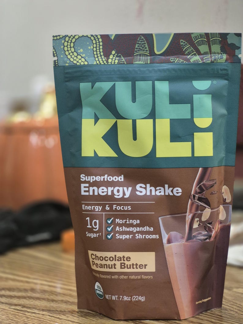 Op de afbeelding: Een verpakking Kuli Kuli Superfood Energy Shake met de smaak Chocolade Pindakaas. De bruin-groene verpakking toont de tekst "KULI". Het bevat 224g product en vermeldt Moringa, Ashwagandha en Super Shrooms als ingredi&euml;nten.