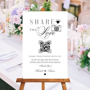 May include: A white sign on a wooden easel with the words "SHARE THE Love" and a QR code. The sign also includes the text "SHARE YOUR PHOTO WITH US!" and "Thank You!" with names below. The background includes floral arrangements.