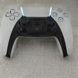 May include: A white and black wireless game controller with a central black panel and two black joysticks. The controller has a light bar and button symbols. The controller is designed for gaming.