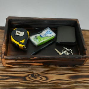 May include: A dark stained wooden tray with various items inside. A yellow and black tape measure, a package of tissues, a black pen, a dark gray wallet, and a set of keys are visible. The tray sits on a wooden surface.