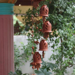 Puede incluir: Un carillón de viento de terracota con una serie de adornos en forma de campana. Las campanas están suspendidas de una pieza superior circular y adornadas con diseños recortados y pequeños elementos decorativos. El carillón de viento cuelga frente a un fondo verde borroso.