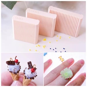 May include: Three light pink rectangular tools with vertical grooves, labeled 1MM, 1.5MM, and 2MM. Below, miniature ice cream cone charms with blue ice cream, chocolate, and cherries. A ring with a green gem and pink accents is also shown.