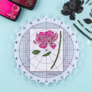 May include: A clear, circular CYFUN DESIGN stamp positioning tool with a white grid and degree markings. A stamped pink flower and green leaves are centered on a white card. Ink pads are visible in the background.