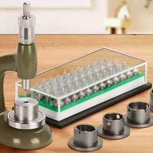 May include: A watch repair tool set, featuring a green press, a clear case with various tips, and several silver metal dies. The tools are arranged on a wooden surface, suggesting a professional or hobbyist setting.