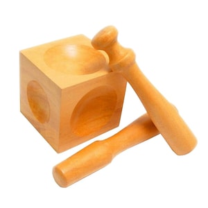 May include: A wooden toy set featuring a cube with rounded indentations and two wooden mallets. The cube has circular cutouts on its sides. The mallets are light brown and have a rounded head and handle.