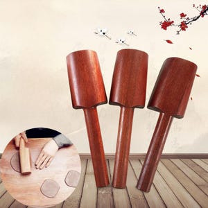 May include: Three wooden rolling pins with a dark reddish-brown finish. The rolling pins have a cylindrical handle and a rectangular head. The background features a light beige wall with decorative elements.