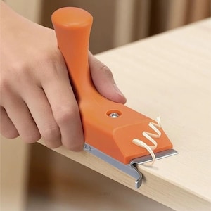 May include: An orange edge scraper tool with a silver blade is used on a wooden surface. The tool has a handle and is removing a thin strip of wood. The number "2.0" is printed on the tool.