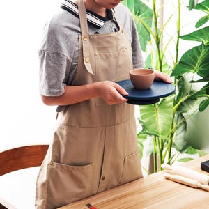 Puede incluir: Delantal color beige con múltiples bolsillos y remaches color latón. Una persona sostiene un torno de alfarero azul oscuro con un pequeño cuenco de cerámica marrón encima. La escena está ambientada en un estudio de cerámica.