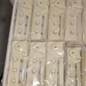 May include: A box filled with multiple off-white electronic devices, each individually wrapped in clear plastic. The devices have a rectangular shape with rounded edges, featuring several circular buttons and a small display area.