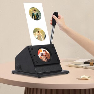 May include: A black photo booth machine with a handle, displaying three oval-shaped images. The images feature illustrations of couples, including a bride and groom. The machine is on a light pink table.