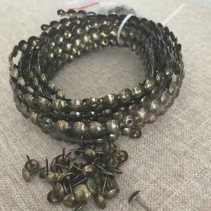 May include: A coil of antique brass upholstery tacks with a decorative scalloped edge. A small pile of individual tacks is also visible. The tacks are a dark, aged brass color, suggesting vintage or rustic decor.