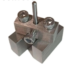 May include: A close-up of a metal tool with a silver finish. The tool is composed of several rectangular blocks and cylindrical components, including three hex-head screws and a central pin. The tool is designed for precision work.