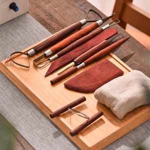 May include: A set of pottery tools arranged on a bamboo tray. The tools include various wooden handled sculpting tools, a wooden paddle, and a block of clay. The tools are a mix of metal and wood.