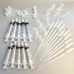 May include: An assortment of clear plastic laboratory supplies, including syringes, funnels, droppers, and stirring rods. The syringes are marked with measurements up to 5ml. The items are arranged on a light-colored surface.