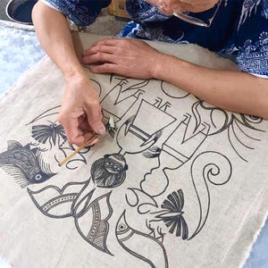 May include: An artisan is shown working on a linen textile, meticulously adding details to a black ink drawing. The artwork features stylized figures, fish, and butterfly motifs, showcasing a blend of artistic skill and cultural design.