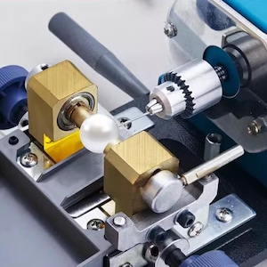 May include: A close-up of a pearl drilling machine. The machine has a blue motor housing, a silver drill bit, and a pearl held in place by two brass-colored clamps. A dark gray handle is visible.
