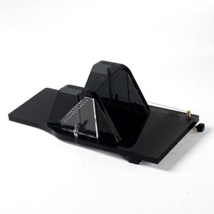 May include: A black acrylic paper cutter with a unique geometric design. It features a triangular blade guide, a curved cutting area, and a thin wire for precise cuts. The base is rectangular with a small knob.